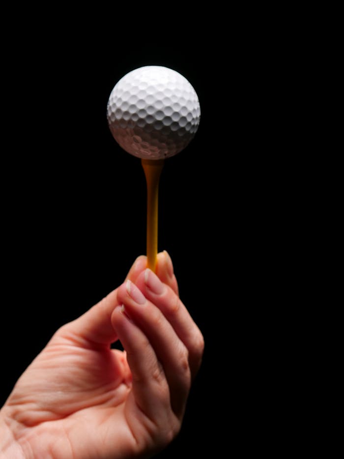 A close-up shot of a hand holding a golf ball on a tee against a black background.